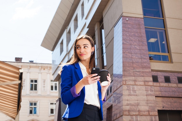 Girl in bright blue jacket stands with smartphone and coffee on the street