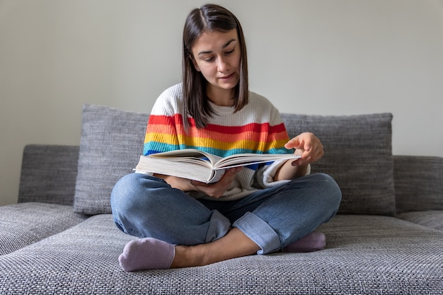밝은 색의 스웨터를 입은 소녀가 집에서 소파에서 책을 읽습니다.