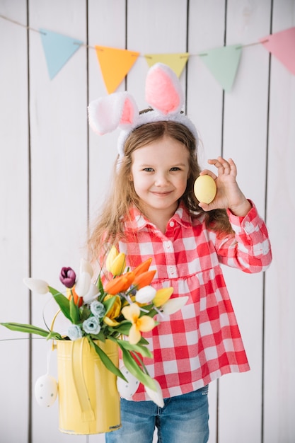 Ragazza nelle orecchie del coniglietto che tiene l'uovo di pasqua e fiori in annaffiatoio