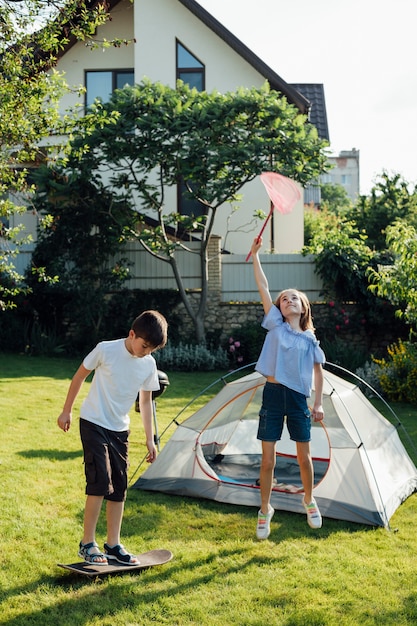 Girl catching butterflies with scoop net and boy playing skateboard near tent camp