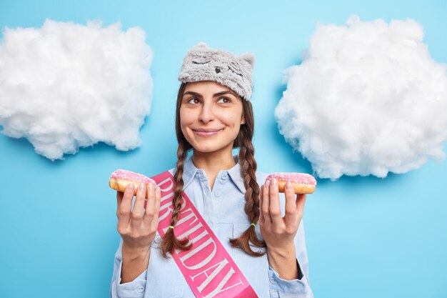 girl celebrates birthday holds two glazed doughnuts dreams about pleasant things dressed in casual clothes isolated on blue 