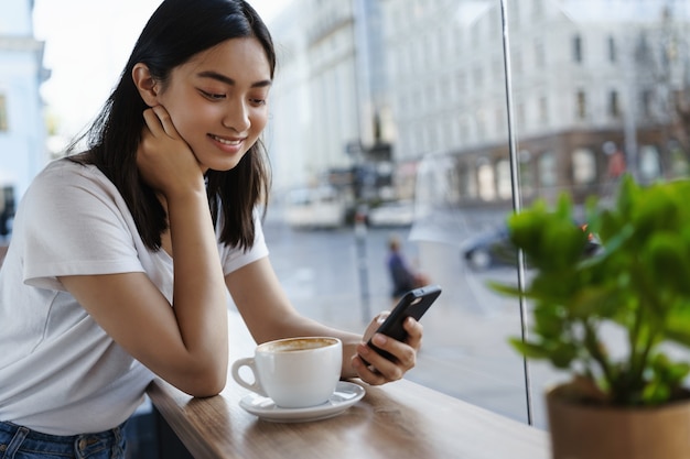スマートフォンでおしゃべりし、窓際のレストランでコーヒーを飲みながら、携帯電話の画面に笑みを浮かべて女の子