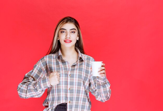 Girl in checked shirt holding a white disposable coffee cup and showing her power