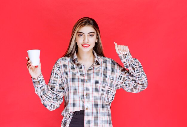 Девушка в клетчатой рубашке держит белую одноразовую кофейную чашку и демонстрирует свою силу