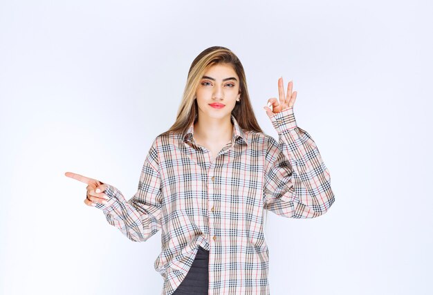 Girl in checked shirt pointing at someone around and showing ok hand sign