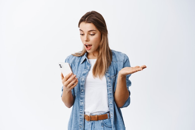 Ragazza che controlla smartphone, guarda lo schermo con una faccia soddisfatta, muro bianco