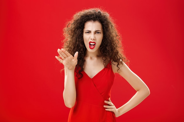 Girl chowing her temper to employees being fed up with unappropriate behaviour complaining standing pissed and dissatisfied gesturing with palm and holding hand on hip over red background.