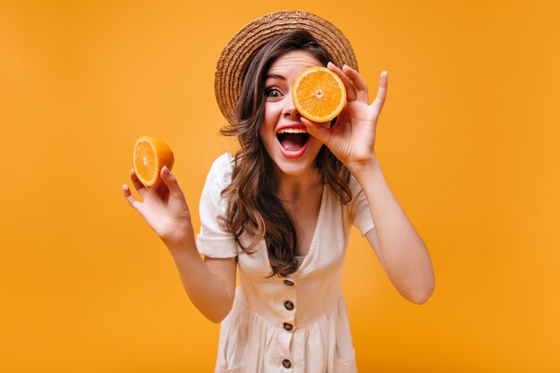 Girl in cotton dress and straw hat is having fun and posing with oranges on isolated background.