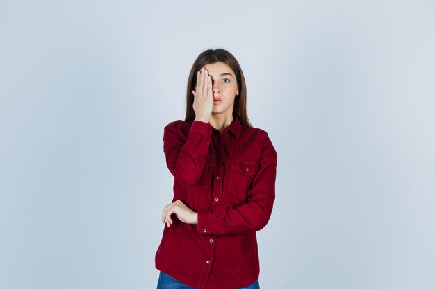 girl covering eye with hand in burgundy shirt and looking wondered.