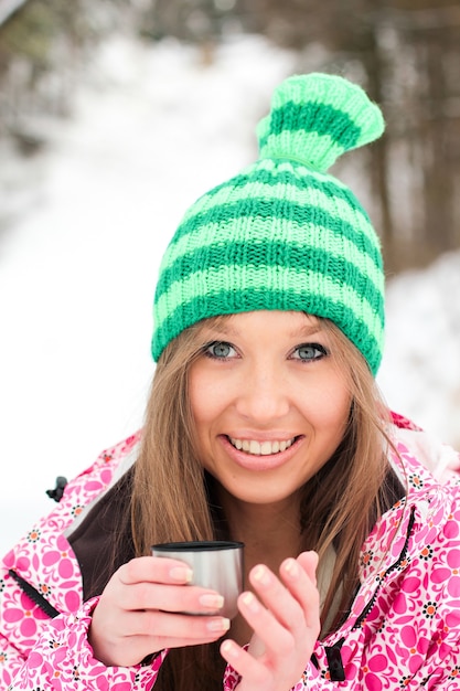 Girl in a crimson jacket and green hat drinking hot tea from a thermos in snowy mountains