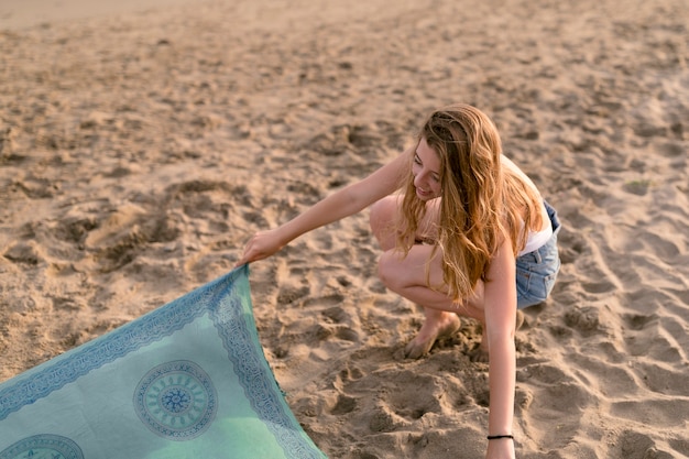 Free photo girl crouching on sand placing scarf at beach