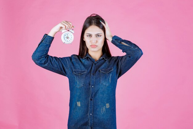Девушка в джинсовой рубашке держит будильник и прикрывает ухо из-за кольца