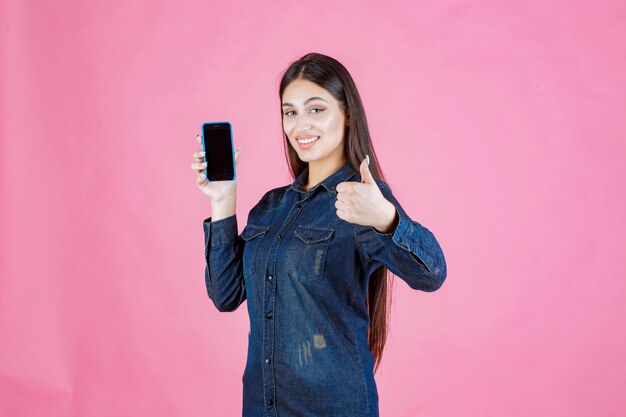 Girl in denim shirt showing her smartphone and making thumb up