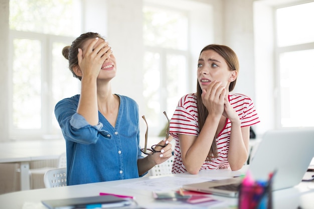 Girl in denim shirt with eyeglasses sadly covering face with hand while upset girl near looking at her Young women working together in modern office