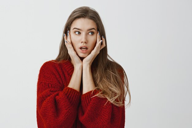 Girl did not know what to do after shocking revelation. Indoor shot of worried attractive caucasian woman in stylish red sweater, holding palms on face and looking aside with troubled, confused face