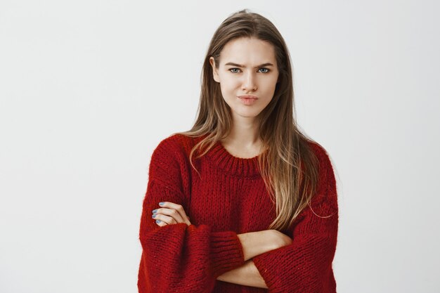 Girl does not buy stupid explanations. Portrait of doubtful displeased creative boss in red loose sweater, holding hands crossed on chest and frowning with smirk, expressing disbelief and hesitation