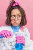Бесплатное фото Девушка проводит исследования с химической жидкостью, работает в лаборатории с образцами жидкости, носит круглые очки и позирует в медицинском халате на розовом