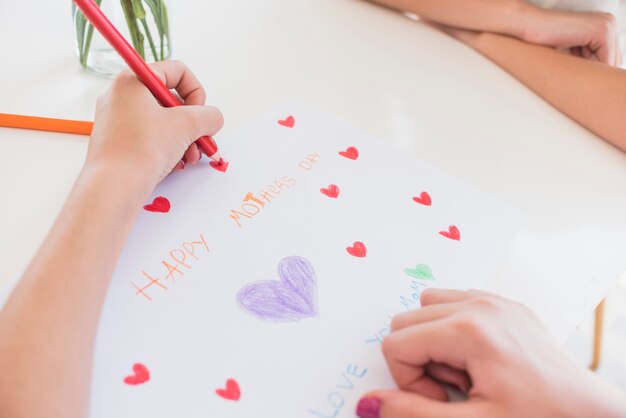 Girl drawing red hearts on paper with Happy Mothers Day inscription 