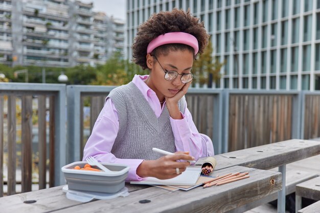 девушка рисует картинки в блокноте, использует цветные карандаши, держит ручки, сосредоточенные в блокноте, носит круглые очки и аккуратную одежду, позирует на улице на фоне городской среды