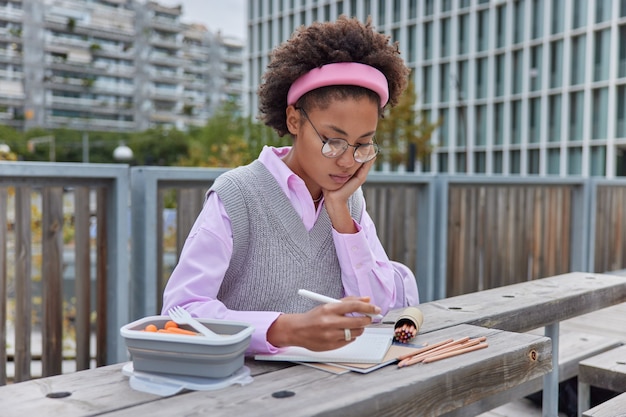 girl draws pictures in notebook uses crayons holds pens concentrated in notepad wears round spectacles and neat clothes poses outside against urban setting