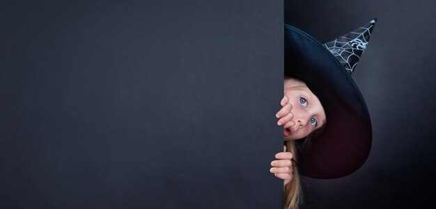 Girl dressed as a witch looks out from behind the wall on a black background banner with copy space