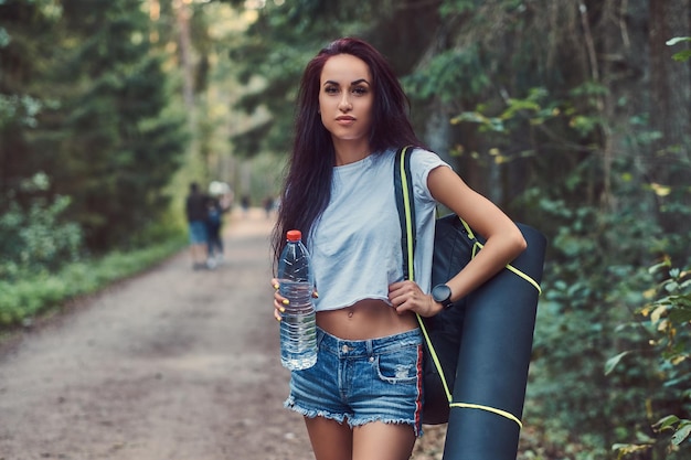 Девушка в рубашке и шортах с туристическим ковриком и рюкзаком держит бутылку воды и смотрит в камеру, стоя в летнем лесу.