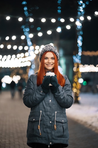 Девушка пьет горячий кофе во время прогулки по вечернему городскому рынку, украшенному праздничными огнями вечером. Ощущение счастья в большом городе. Проводим зимние каникулы