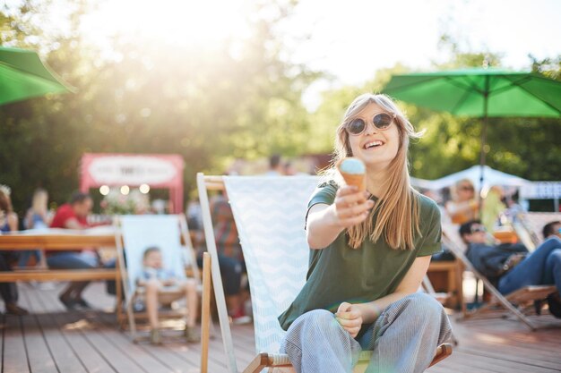 웃고 있는 아이스크림을 먹는 소녀 카메라에 제안하는 여름을 즐기는 안경을 쓰고 카메라를 쳐다보며 아이스크림을 먹고 있는 화창한 날 공원에 앉아 있는 젊은 여성의 초상화 여름 라이프스타일 컨셉