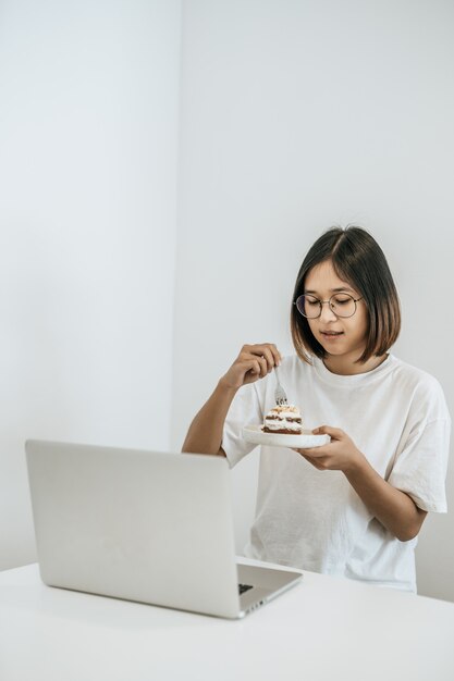 女の子はケーキを食べ、テーブルの上にラップトップを持っています。