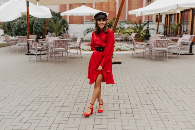 Foto gratuita la ragazza in vestito elegante e rosso con cintura e scarpe sul tallone della città mostra le gambe snelle. foto a figura intera nel caffè della città