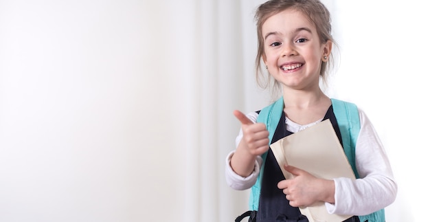 Foto gratuita ragazza-studentessa di scuola elementare con uno zaino e un libro su uno sfondo chiaro. il concetto di istruzione e scuola primaria. posto per il testo.