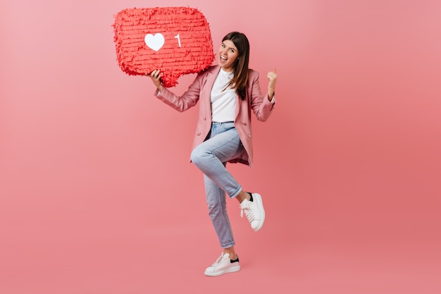 Girl enjoying social network feedback. Studio shot of young woman dancing with like icon on pink background.