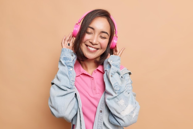 Girl enjoys music catches every bit of song wears wireless pink headphones denim jacket closes eyes from satisfactio poses on brown likes playlist