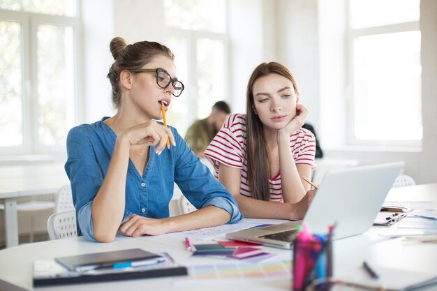 Girl in eyeglasses with pencil and thoughtful girl in striped Tshirt leaning on hand while thoughtfully working with laptop together Young women spending time in modern office