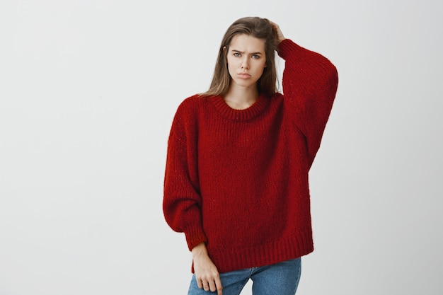 Girl feeling bothered not knowing answer to question. Portrait of displeased gloomy european woman in trendy loose sweater, scratching back of head and frowning, confused and troubled over gray wall