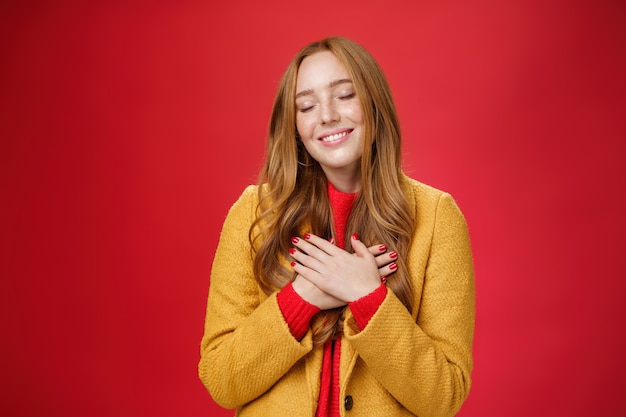 Ragazza che si sente come il riscaldamento del cuore. come ricordare dolci teneri ricordi con gli occhi chiusi e un ampio sorriso sciocco tenendosi per mano sul petto premendo sull'anima in posa nostalgica e commossa su sfondo rosso.