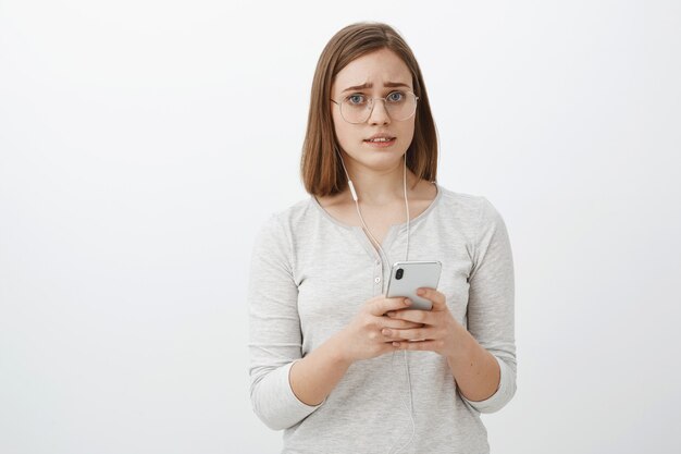 Girl feeling worried one earphone broke down. Portrait of displeased and concerned upset cute woman in glasses with short brown hair frowning and making sad face holding smartphone wearing headphones