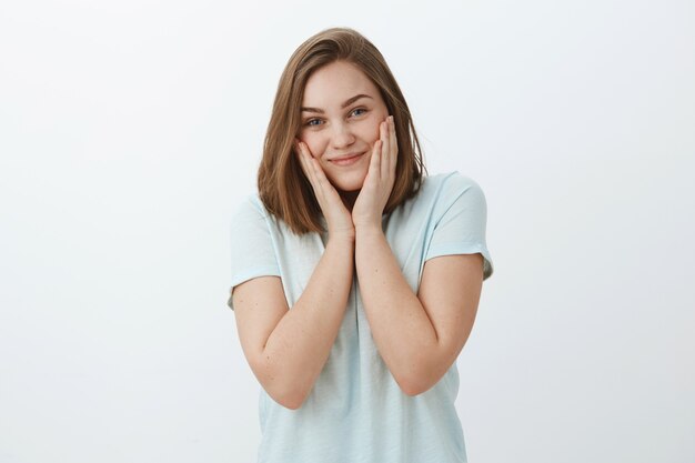 Girl finally get rid of acne with new facial mask. Charming pleased and bright woman touching cheeks and smiling joyfully feeling beautiful and fresh solving skin problems posing against grey wall