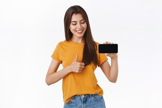 Girl fully satisfied with awesome new mobile application recommend everyone download Pleased goodlooking woman in yellow tshirt holding smartphone horizontally showing thumbsup