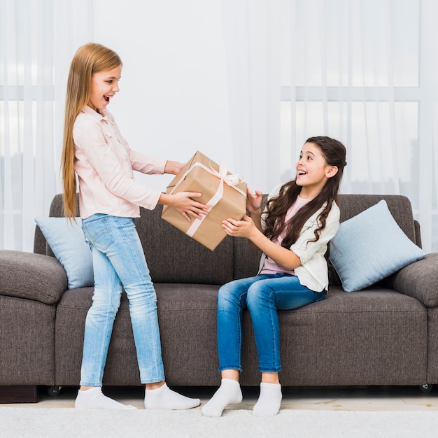 Girl giving present to her surprised friend sitting on sofa in the living room