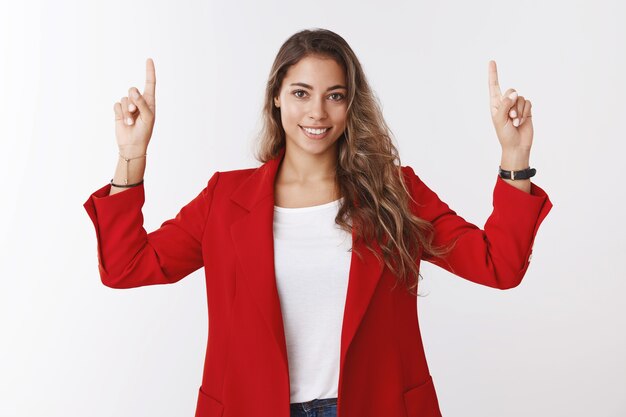 Цель девушки занять первое место. Уверенная в себе и решительная великолепная женщина-предприниматель 25-х годов в красной куртке поднимает руки, указывая указательным пальцем вверх, показывая пространство для копирования, улыбаясь, повезло, уверенно