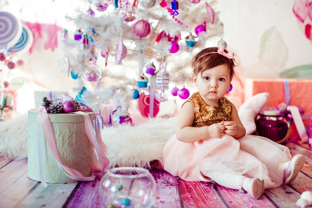 Girl in golden dress sits among present boxes before Christmas tree 