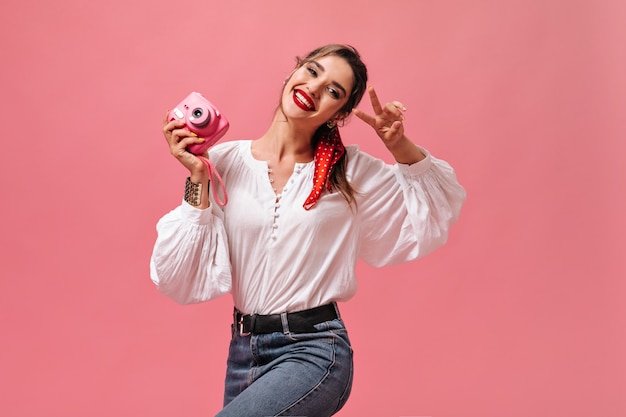 Girl in good mood holds camera and shows peace sign on pink background.  Lovely lady with red lipstick in white blouse and jeans is smiling.