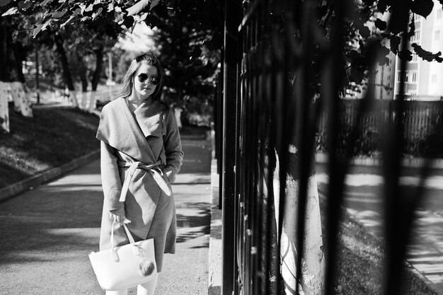 Girl in gray coat with sunglasses and handbag walking at street against iron fence