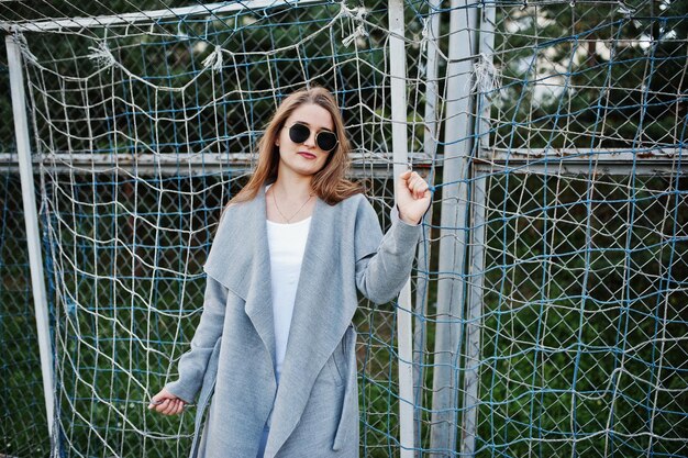 Девушка в сером пальто с солнечными очками у футбольных ворот небольшого уличного стадиона
