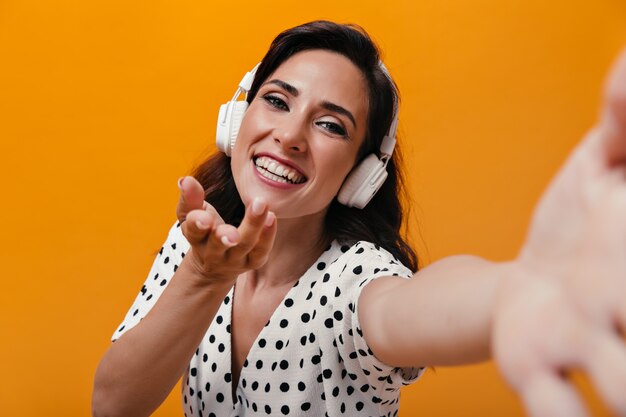 Girl in great mood is listening to music with headphones and taking selfie on orange background