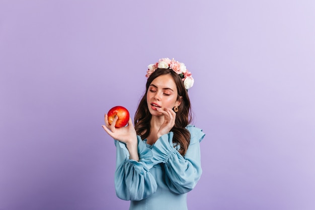 Девушка в прекрасном настроении смотрит на вкусное красное яблоко. Снимок женщины в голубой блузке на изолированной стене.