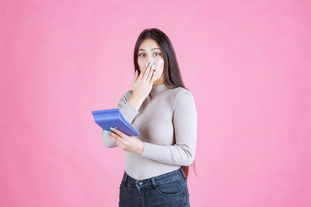 Girl in grey shirt holding a blue calculator and showing silence sign as she is working