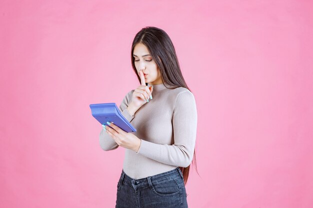 Girl in grey shirt holding a blue calculator and showing silence sign as she is working