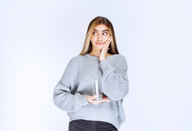 Girl in grey sweatshirt holding a glass of pure water and making doubting face.